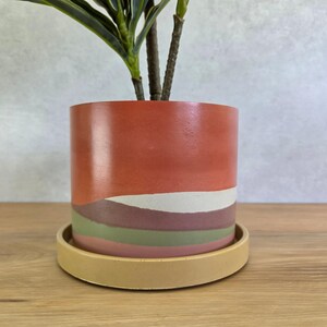 May include: A cylindrical plant pot featuring a smooth finish with horizontal, wavy color bands in shades of terracotta, cream, dark brown, olive green, and dusty pink. The pot sits on a matching circular saucer in a solid, muted mustard yellow.