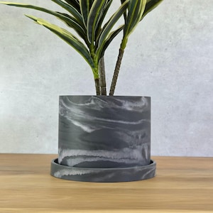 May include: A cylindrical plant pot with a dark charcoal gray and white marbled pattern, featuring a matching circular saucer base. A green and yellow variegated plant with long, pointed leaves is planted inside the container.