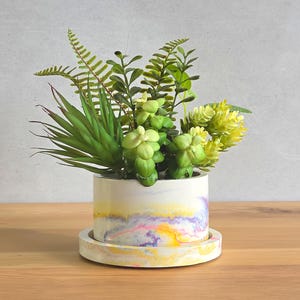 May include: A cylindrical white planter with a marbled pattern of yellow, pink, and purple swirls, resting on a matching circular tray. The planter contains an arrangement of various green artificial ferns and succulent-like plants.
