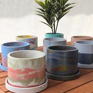 May include: A collection of cylindrical concrete planters with matching saucers, featuring various marbled and solid color patterns including sage green, coral, blue, yellow, and dark grey, arranged on a wooden surface with a green plant.