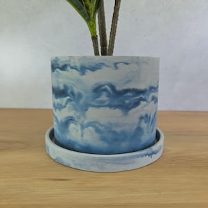 May include: A cylindrical plant pot with a matching circular saucer, featuring a marbled pattern of swirling blue and white tones. The exterior surface displays a cloud-like aesthetic with varying shades of blue blended into a white base.