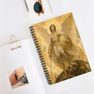 May include: A spiral-bound notebook featuring a golden illustration of an angel with large wings, wearing armour and holding a sword whilst standing over a dark creature. The cover art is rendered in shades of gold and yellow with a radiant halo effect.