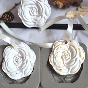 May include: Three white rose-shaped ornaments made of a solid material, each featuring a cream-coloured ribbon loop for hanging. One ornament is plain white, one has red speckles, and one has gold-coloured accents, resting near a grey mould.