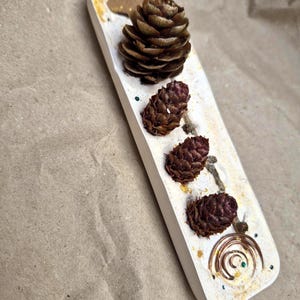 May include: A rectangular white tray featuring four brown pinecones of varying sizes arranged in a line. The tray surface is decorated with gold-coloured flecks, small green accents, a thin twig, and a circular copper wire spiral at the bottom end.