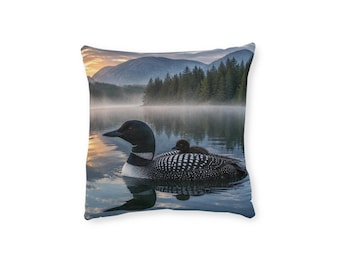 Può includere: Un cuscino quadrato con una stampa fotografica di un adulto di strolaga con un piccolo pulcino sul dorso, che nuota su un lago calmo e nebbioso all'alba. Lo sfondo mostra una fitta foresta di pini e montagne lontane sotto un cielo tenue.