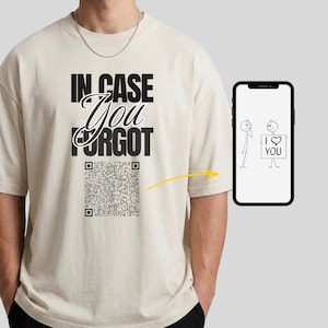 以下が含まれることがあります： ベージュの半袖Tシャツ。黒字で「IN CASE You FORGOT」と書かれ、その下にQRコードが配置されている。黄色い矢印がスマートフォンの画面を指しており、そこには「I LOVE YOU」と書かれた看板を持つ棒人間のイラストが表示されている。