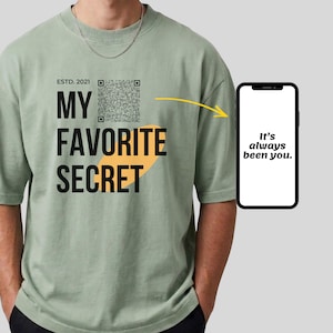 以下が含まれることがあります： セージグリーンのTシャツで、黒い文字で「ESTD. 2021 MY FAVORITE SECRET」とQRコードがプリントされています。黄色い矢印が、白い画面に「It's always been you.」と表示されたスマートフォンを指しています。Tシャツはクルーネックです。