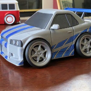 May include: A silver toy car with blue racing stripes on the bonnet and blue graphic decals on the sides. The vehicle features a prominent blue rear spoiler, black windows, and large silver five-spoke wheels with black tyres in a stylised design.