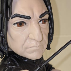 May include: A 3D printed figurine of a person with shoulder-length black hair, brown eyes, and a stern facial expression. The figure wears a black collared garment and holds a thin black wand extended forward in a pointing gesture.