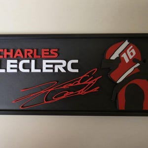 以下が含まれることがあります： 黒い長方形の壁掛け看板。赤い文字で「CHARLES」、白い文字で「LECLERC」と書かれています。名前の下には赤いサインがあります。右側には赤と黒のレーシングヘルメットのグラフィックがあり、白い斜めのストライプの上に白い数字の16が表示されています。