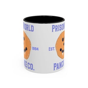 Puede incluir: Una taza de cerámica blanca con interior negro que presenta un gráfico de un panqueque color canela con arándanos oscuros y el texto PRISON WORLD PANCAKE CO. EST. 1994 en letras azules. El diseño está impreso en el exterior de la taza cilíndrica.