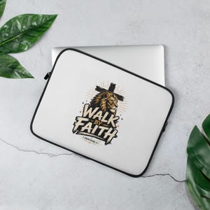 May include: A white laptop sleeve with a black zipper border featuring a graphic of a lion head in front of a cross. The text reads WALK BY FAITH and 2 CORINTHIANS 5:7 in a bold, stylized font. The design includes small star accents and sunburst lines.