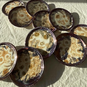 May include: A collection of ten round ceramic plates featuring a marbled glaze pattern in shades of cream, tan, and dark brown. Each plate has a glossy finish with swirling, organic circular motifs that create a unique, fluid appearance.