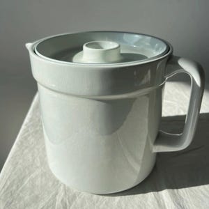 Może przedstawiać: Biały ceramiczny dzbanek o cylindrycznym korpusie, płaskiej pokrywce z centralnym uchwytem w kształcie gałki oraz bocznym uchem. Powierzchnia dzbanka wykazuje subtelny, drobny wzór spękań szkliwa na całej zewnętrznej stronie.