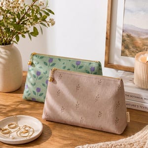 Könnte beinhalten: Zwei rechteckige Reißverschlusstaschen mit goldfarbenen Beschlägen. Eine Tasche ist hellgrün mit einem violetten Tulpenmuster, die andere ist altrosa mit einem kleinen, sich wiederholenden dunkelbraunen Blattdruck und einem Etikett mit der Aufschrift LEAVES.