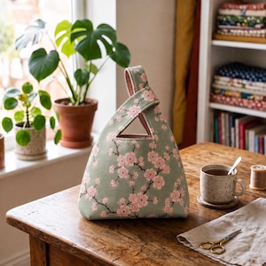 Pode incluir: Uma bolsa de tecido com nó sobre um fundo verde sálvia claro com um padrão repetitivo de ramos de cerejeira em flor cor-de-rosa. A bolsa tem uma forma triangular com uma alça integrada única e um forro interior rosa suave.