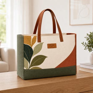 Può includere: Una borsa tote rettangolare con corpo in tela color crema e base e pannelli laterali verde scuro. La parte anteriore presenta un motivo a foglie stampato in tonalità di verde e oro, un accento curvo color ruggine e un'etichetta KORA.