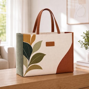 May include: A rectangular tote bag with two brown handles, featuring a cream-colored front panel with a printed leaf design in shades of green and mustard yellow. The side panel is olive green, and the right side is rust orange with a brown KORA label.