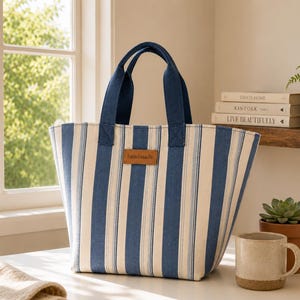Puede incluir: Un bolso tote con rayas verticales en tonos azul marino, azul claro y blanco roto. El bolso tiene dos asas de tela azul oscuro y un parche rectangular de color marrón similar al cuero en el centro con la palabra handmade grabada.