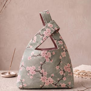 Pode incluir: Uma bolsa de tecido tipo nó com fundo verde sálvia e um padrão repetitivo de flores de cerejeira cor-de-rosa e ramos castanhos escuros. A bolsa tem uma forma triangular com uma alça integrada e um forro interior em tom malva sólido.