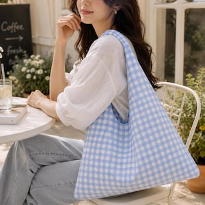 May include: A light blue and white gingham patterned hobo-style shoulder bag made of fabric. The bag features a soft, unstructured silhouette with a wide integrated strap, showcasing a classic checkered print throughout the entire exterior surface.