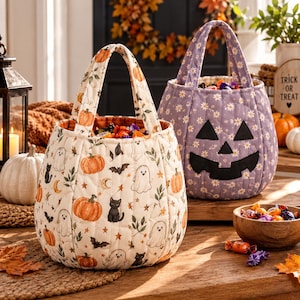 May include: Two quilted fabric trick-or-treat bags. One bag is cream-colored with a pattern of pumpkins, ghosts, cats, and bats. The other bag is light purple with a white daisy print and a black fabric jack-o'-lantern face appliqué.