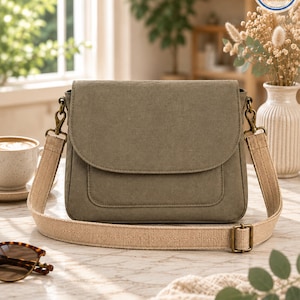 May include: An olive green canvas crossbody bag with a fold-over flap and a front pocket. The bag features a wide, adjustable beige woven strap attached with antique brass-toned metal hardware clips and a sliding buckle for length adjustment.