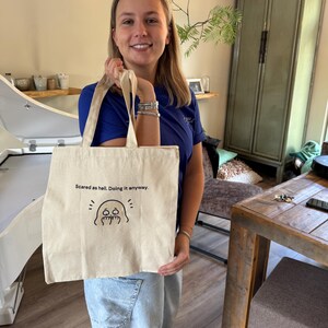 Op de afbeelding: Een beige canvas draagtas met zwarte tekst die luidt "Scared as hell. Doing it anyway." en een eenvoudige zwarte lijntekening van een spookachtig figuurtje met grote ogen en een golvende mond onder de tekst.