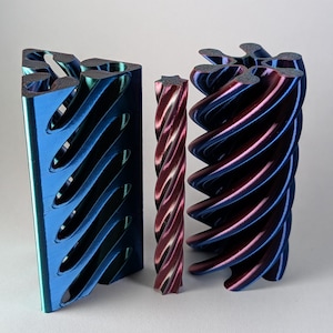 May include: Three 3D printed objects with a metallic finish. On the left, a triangular prism with diagonal cutouts. In the center, a thin, spiraled rod. On the right, a cylindrical object with a deep, spiraled, multi-faceted geometric design.