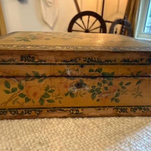 May include: A rectangular wooden box with a distressed yellow finish, featuring hand-painted floral motifs in shades of orange and green. The box is decorated with a dark green ornate border pattern and includes a central metal keyhole on the front.