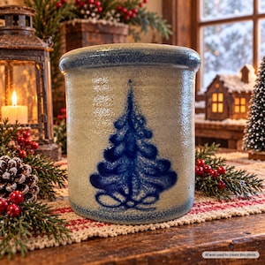 May include: A cylindrical stoneware crock with a speckled light gray finish and a thick, rounded rim. The front features a hand-painted cobalt blue evergreen tree design, with a matching blue band around the top and bottom edges.