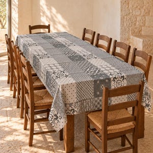 May include: A rectangular tablecloth featuring a patchwork pattern of various black, white, and grey geometric and floral motifs. The tablecloth is draped over a long wooden table surrounded by eight wooden chairs with woven straw seats.