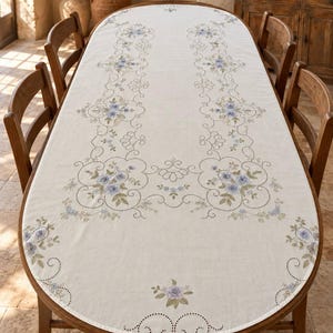 May include: An oval-shaped off-white tablecloth featuring intricate cutwork embroidery and delicate floral motifs in shades of blue, lavender, and sage green. The design includes symmetrical scrolling patterns and detailed flower clusters.