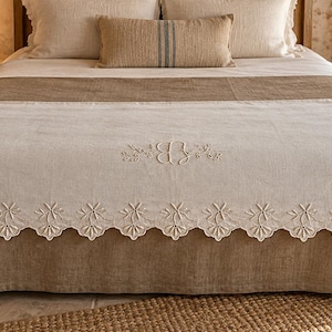 Puede incluir: Una colcha de color beige claro con un borde festoneado blanco que presenta un intrincado bordado floral calado. El centro de la colcha muestra un delicado monograma bordado con la letra B rodeada de pequeñas ramas y enredaderas.