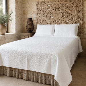 Puede incluir: Una colcha blanca con un patrón floral texturizado que cubre una cama con dos almohadas blancas a juego. La cama cuenta con un volante de ganchillo beige y está colocada contra un gran cabecero de madera tallada en marrón claro.