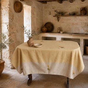 Puede incluir: Un mantel rectangular con un diseño bicolor que presenta una sección superior en amarillo mostaza y un borde inferior en beige claro. La tela está decorada con pequeñas ramas florales bordadas en verde y amarillo sobre la superficie.