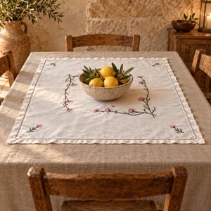 Pode incluir: Uma toalha de mesa quadrada de linho em tom off-white com uma delicada borda bordada de caules verdes e pequenas flores cor-de-rosa. Centralizada no tecido está uma tigela de cerâmica salpicada cheia de três limões amarelos e folhas.