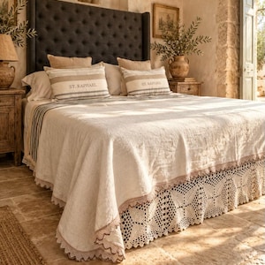 Puede incluir: Una cama con un cabecero alto capitoné de color gris oscuro, una colcha texturizada de color crema con un borde de encaje festoneado y dos almohadas rectangulares con rayas horizontales y el texto ST. RAPHAEL impreso en el centro de cada una.