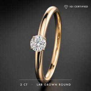 Puede incluir: Un anillo de oro con un diamante redondo cultivado en laboratorio en un engaste de cuatro garras. La banda es lisa y pulida. El texto en la imagen dice IGI CERTIFIED y 2 CT LAB GROWN ROUND, indicando el peso y la certificación del diamante.