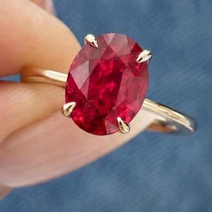 Op de afbeelding: Een ring met een grote, ovaal geslepen, dieprode edelsteen gezet in een gepolijste gouden band. De steen wordt vastgehouden door vier gouden pootjes en heeft een gefacetteerd oppervlak dat licht reflecteert. De band is dun, glad en van goudkleurig metaal.