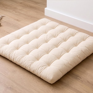 May include: A square, off-white floor cushion featuring a tufted design with multiple indentations across its surface. The cushion has a low-profile, padded construction and a soft, fabric exterior, providing a flat, cushioned surface for floor seating.