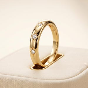 May include: A polished gold band ring featuring three evenly spaced, flush-set clear stones. Each stone is centered within a star-shaped engraving etched directly into the smooth, rounded surface of the gold metal band.