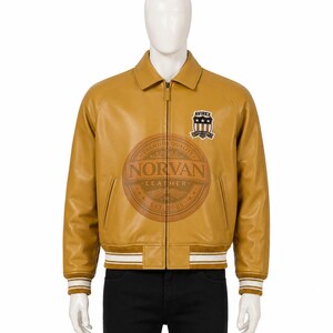 K&ouml;nnte beinhalten: Eine senfgelbe Lederjacke mit Frontrei&szlig;verschluss, klassischem Kragen sowie gerippten B&uuml;ndchen und Saum mit wei&szlig;en Streifen. Auf der Brust befindet sich ein Avirex-Aufn&auml;her mit Sternen und Streifen. Ein kreisf&ouml;rmiges Norvan Leather Logo ist &uuml;berlagert.