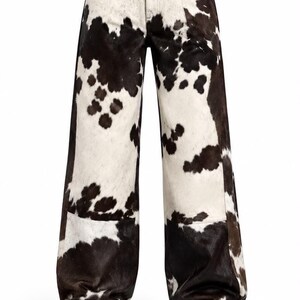 Könnte beinhalten: Lange Hose mit einem schwarz-weißen Kuhfellmuster. Die Hose verfügt über einen Knopf- und Reißverschluss an der Taille und einen geradlinigen Schnitt. Ein runder brauner Lederaufnäher in der oberen rechten Ecke zeigt den Text NORVAN LEATHER.