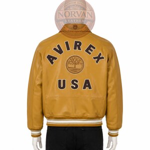 K&ouml;nnte beinhalten: Eine senfgelbe Leder-Collegejacke mit dem Schriftzug AVIREX in dunkelbraunen Buchstaben, die &uuml;ber einem kreisf&ouml;rmigen Baumlogo gebogen sind, und dem Schriftzug USA darunter. Die Jacke hat einen gerippten Kragen, B&uuml;ndchen und Saum mit wei&szlig;en Streifen.