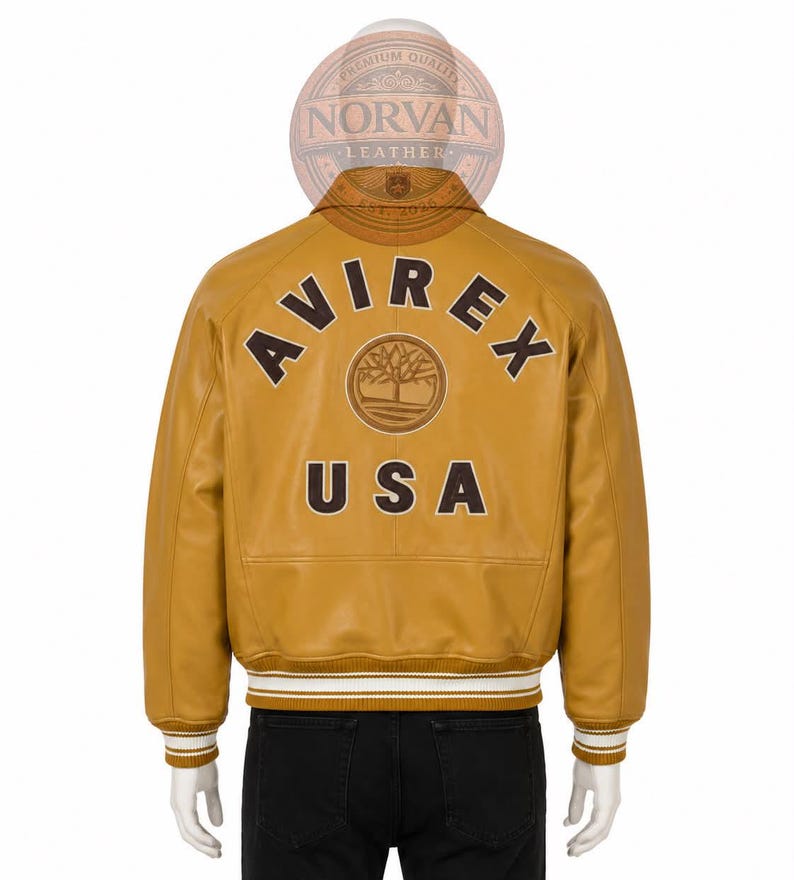 K&ouml;nnte beinhalten: Eine senfgelbe Leder-Collegejacke mit dem Schriftzug AVIREX in dunkelbraunen Buchstaben, die &uuml;ber einem kreisf&ouml;rmigen Baumlogo gebogen sind, und dem Schriftzug USA darunter. Die Jacke hat einen gerippten Kragen, B&uuml;ndchen und Saum mit wei&szlig;en Streifen.