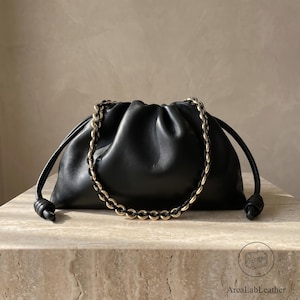 May include: A black leather pouch bag with a gathered, soft silhouette and a drawstring closure. The bag features a decorative chain strap with alternating gold-tone metal links and black leather accents, creating a structured contrast.