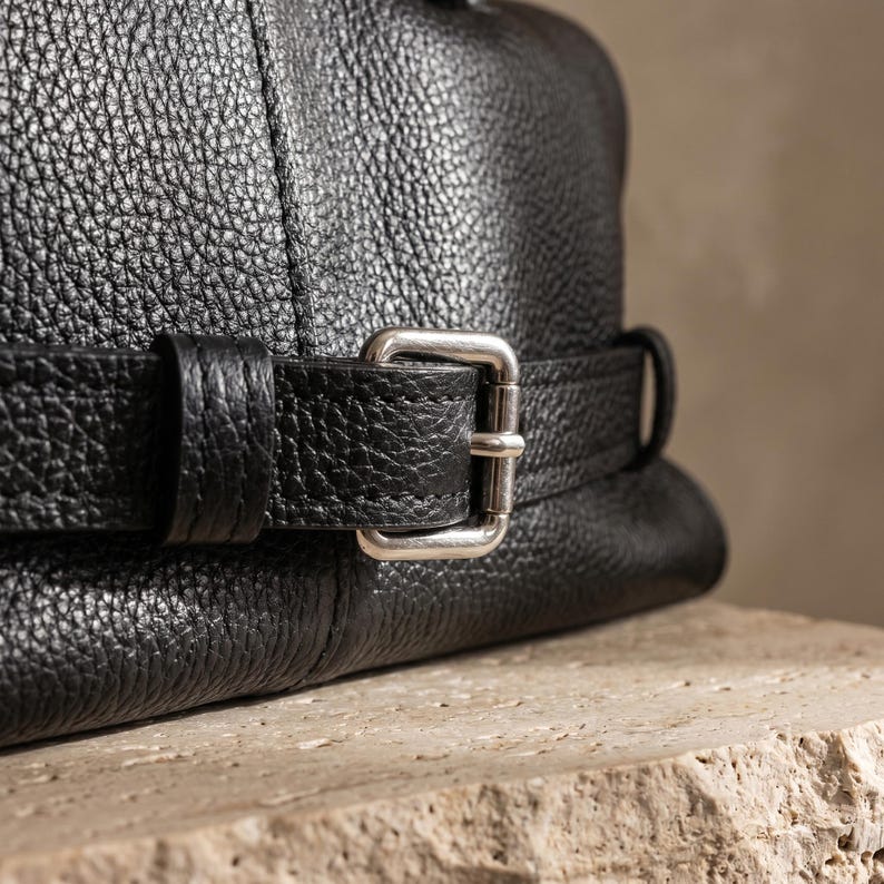 Close-up of the silver buckle and leather belt strap on the front of the black genuine leather office handbag, the design detail used to adjust the bag's volume.
