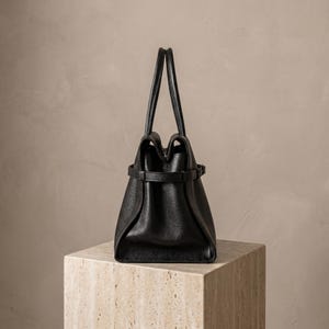 Side profile of a structured black leather office handbag and crossbody purse, showing depth, rolled top handles and the thin adjustable shoulder strap for crossbody wear.