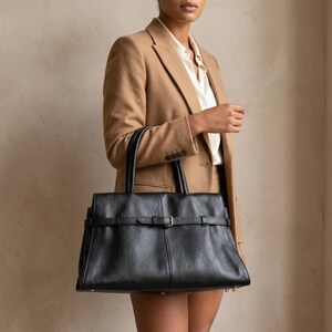 Lifestyle torso shot of a woman carrying the large office handbag by its top handles, the textured black genuine leather purse styled with a polished quiet-luxury work outfit.